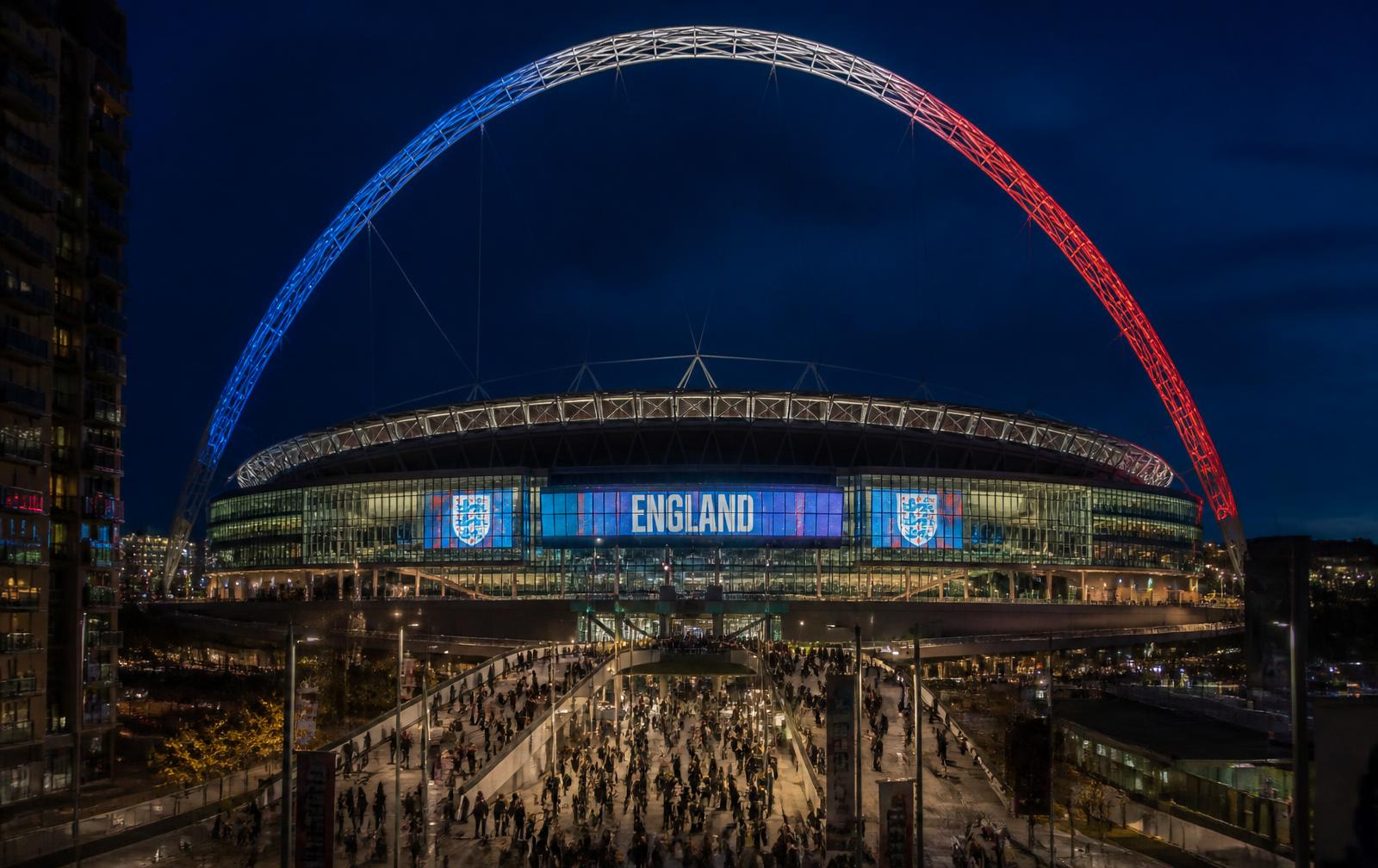 Floodlit football stadium glowing at night above arriving crowds