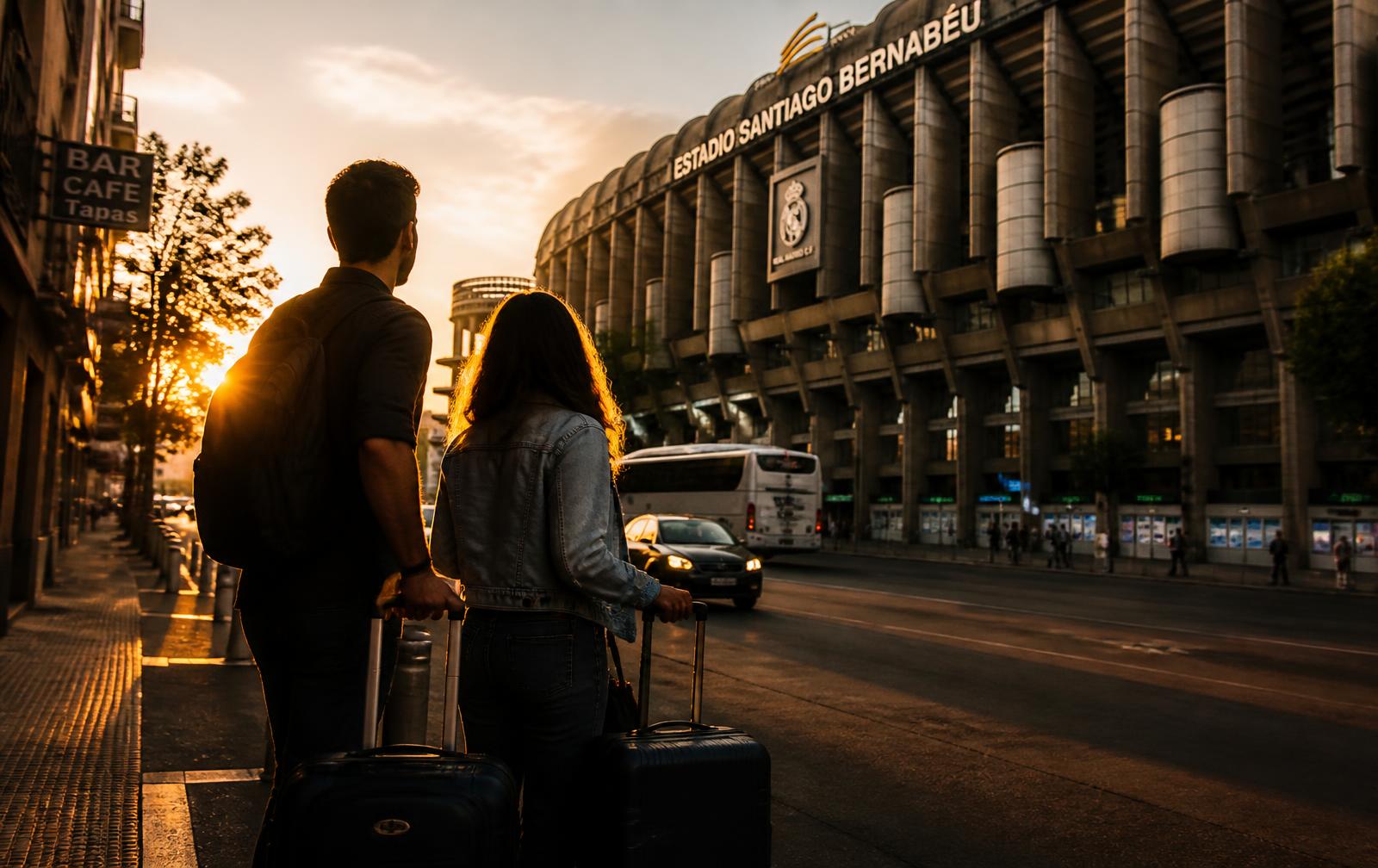 Two travellers with luggage arriving at a stadium in a European city at sunset